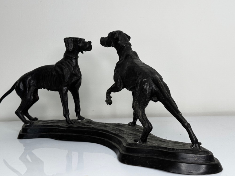 A dark patinated bronze sculpture depicting two hunting hound dogs in active poses on an integral naturalistic base. View XXV