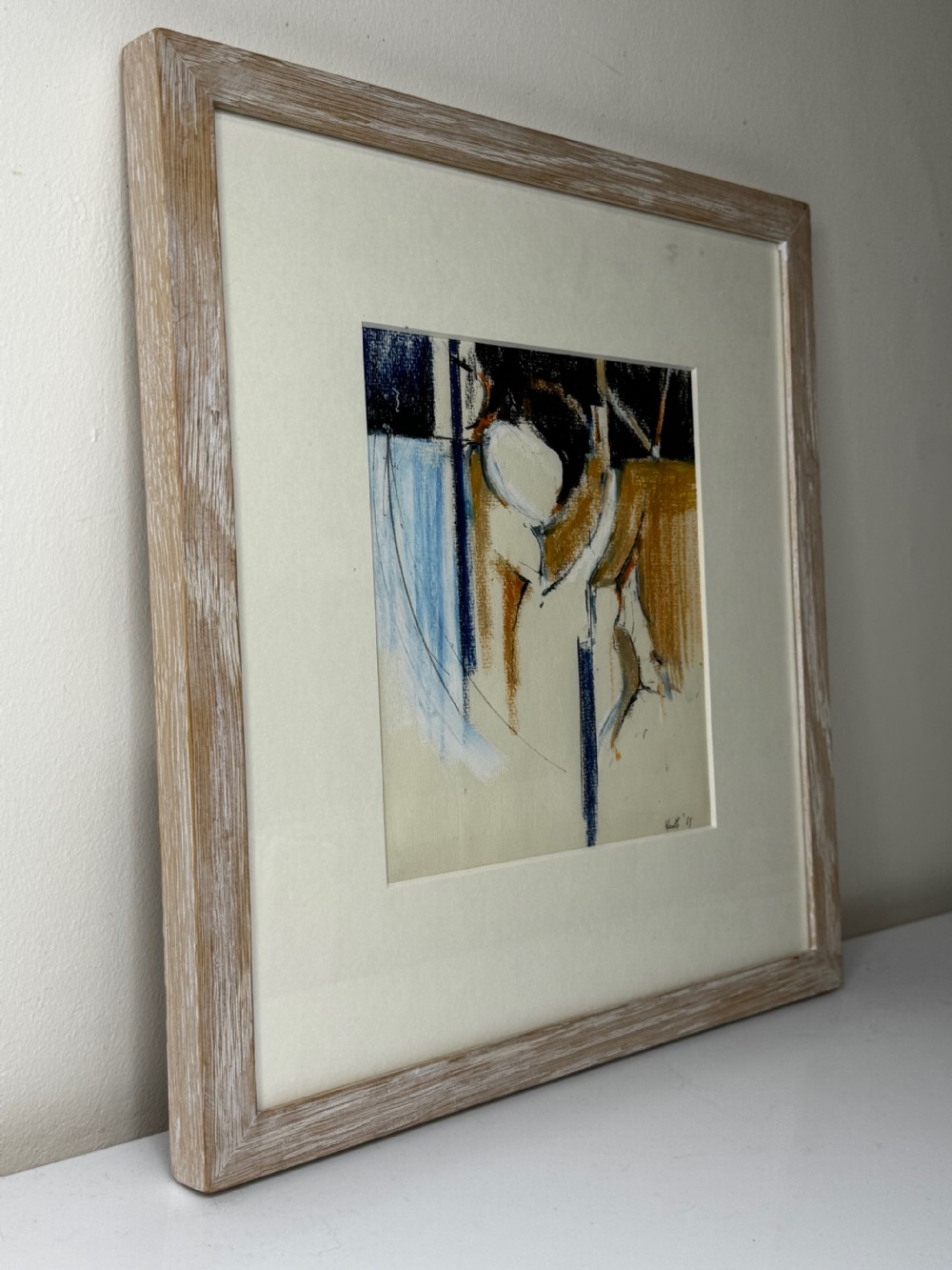 A framed abstract watercolour and pastel painting featuring gestural shapes in ochre, navy blue, and cream enclosed within a wide light wood frame. View V