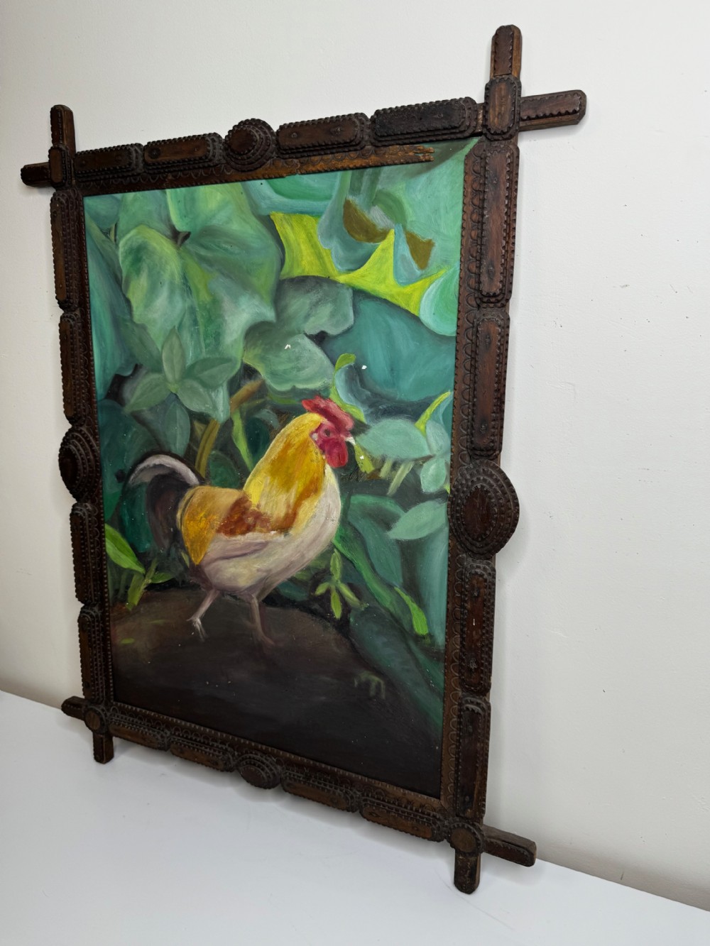 An oil painting depicting a yellow and red cockerel amidst dense green leaves, set within a carved dark wood frame with extended corner joints. View X