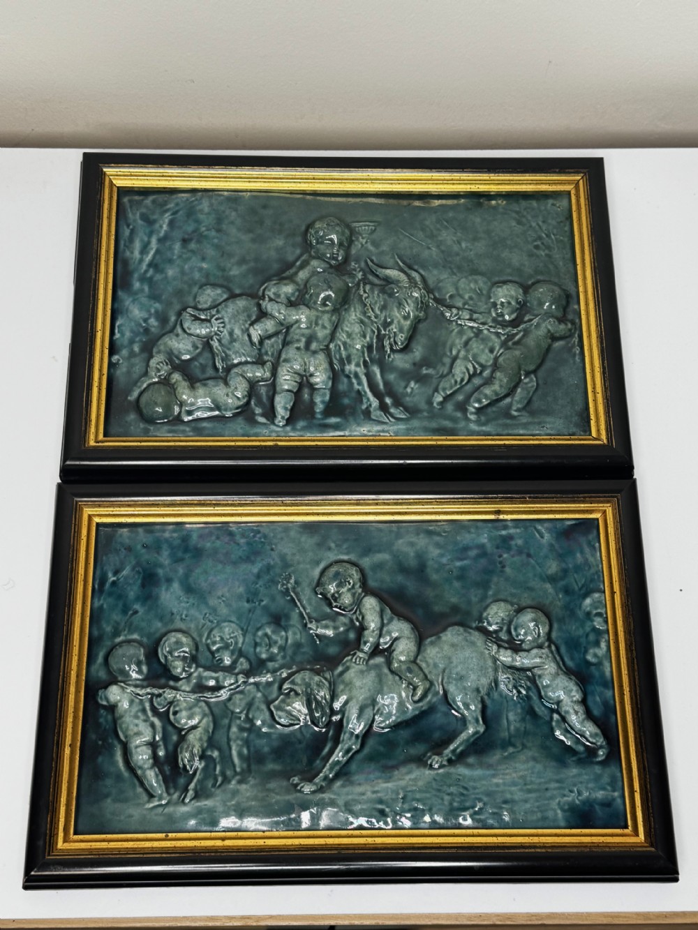 A pair of rectangular Arts and Crafts earthenware relief plaques with turquoise glaze displaying scenes of children with a goat and dog set in ebonised frames. View VI