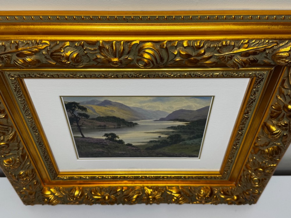 An early 20th-century framed watercolour painting depicting a serene Lake District landscape with a body of water and mountains, framed in a heavy gilt floral moulding. View VI