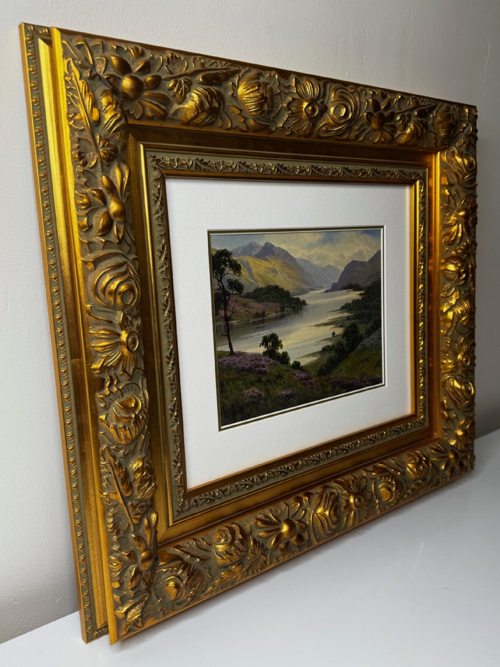 An early 20th-century framed watercolour painting depicting a serene Lake District landscape with a body of water and mountains, framed in a heavy gilt floral moulding. View VII