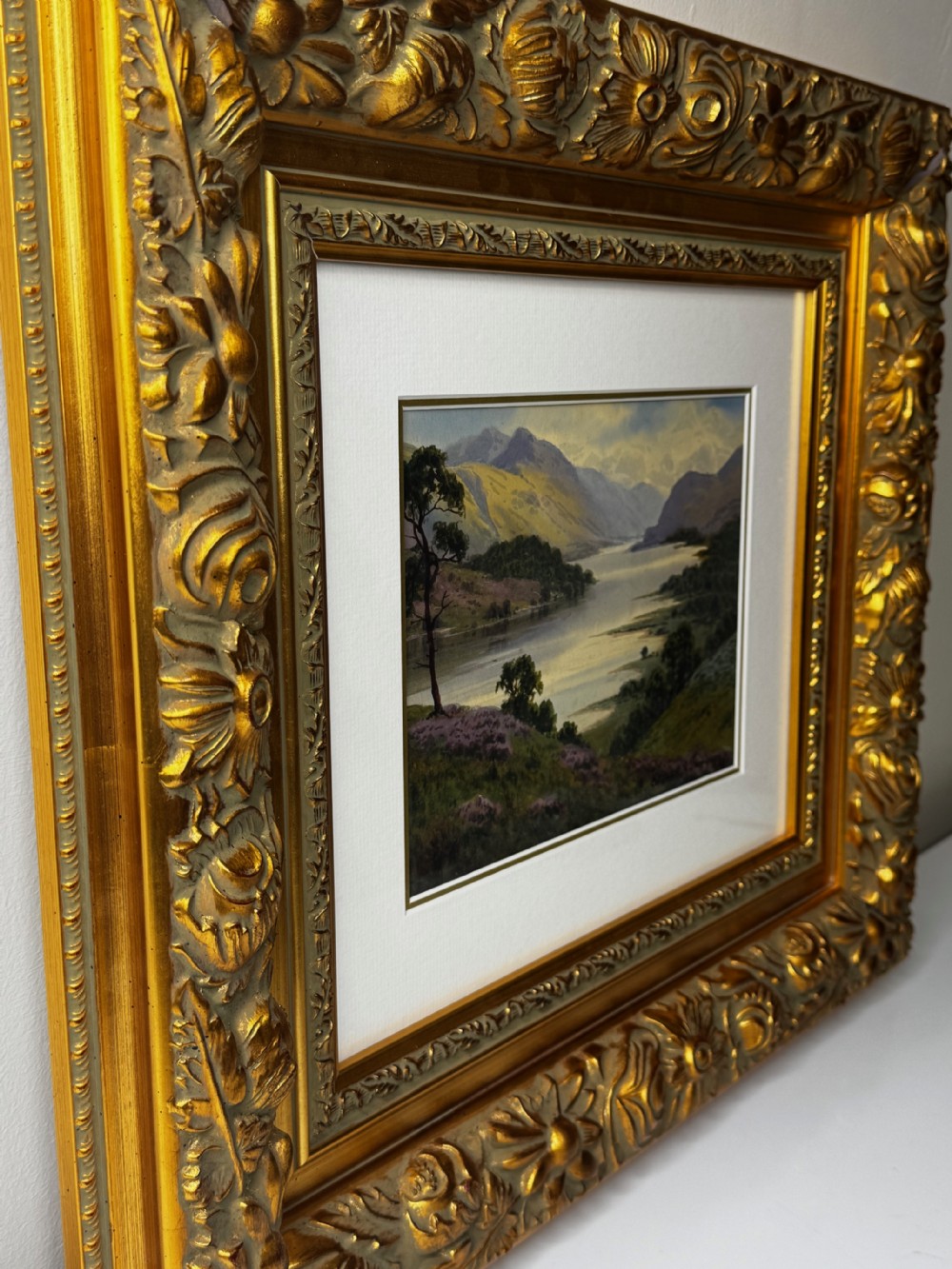An early 20th-century framed watercolour painting depicting a serene Lake District landscape with a body of water and mountains, framed in a heavy gilt floral moulding. View VIII