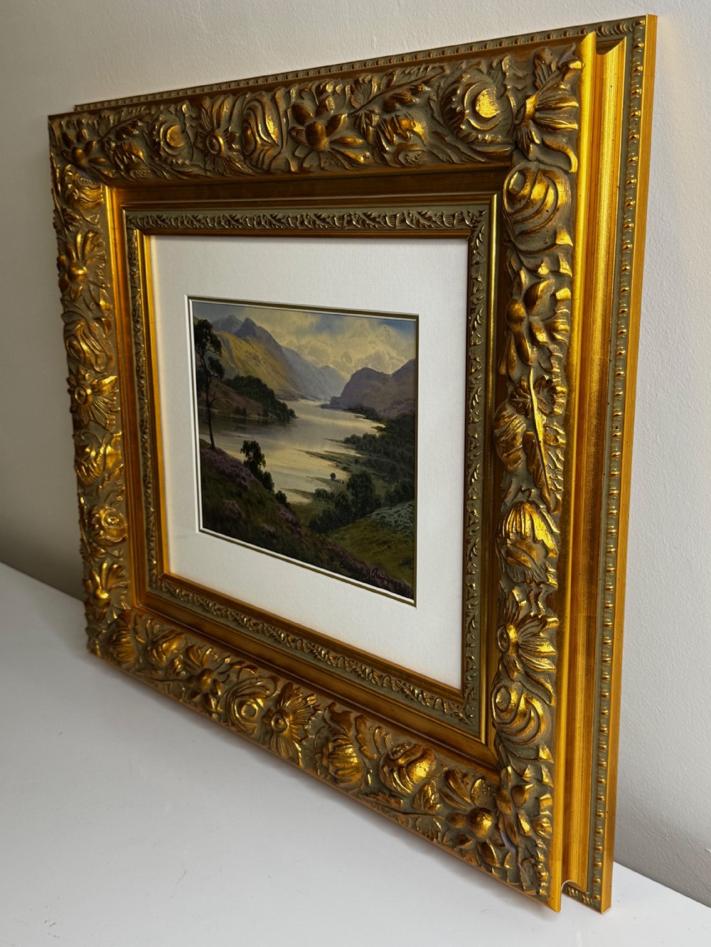 An early 20th-century framed watercolour painting depicting a serene Lake District landscape with a body of water and mountains, framed in a heavy gilt floral moulding. View X