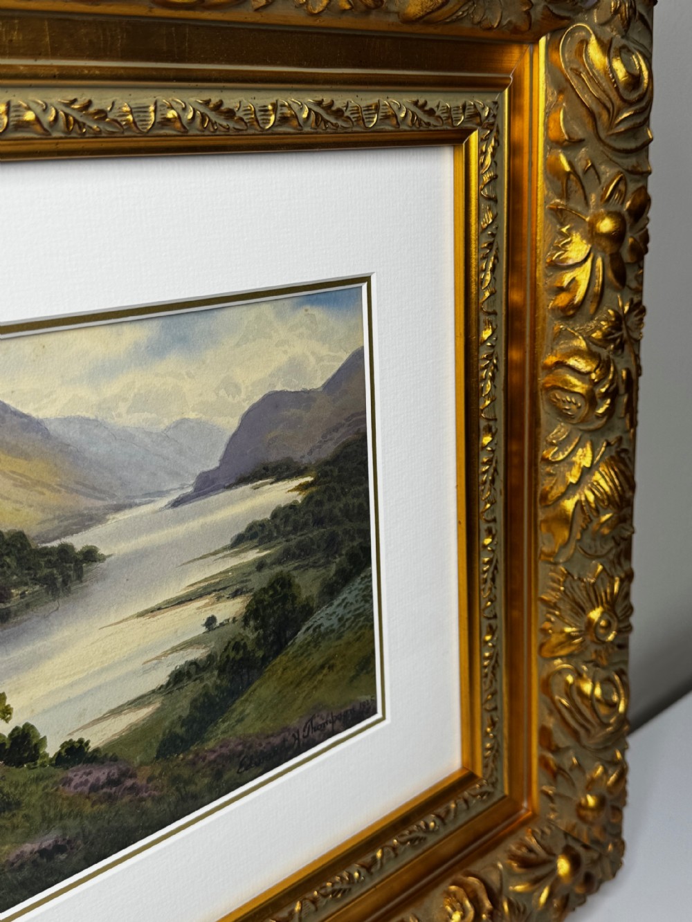 An early 20th-century framed watercolour painting depicting a serene Lake District landscape with a body of water and mountains, framed in a heavy gilt floral moulding. View XIII