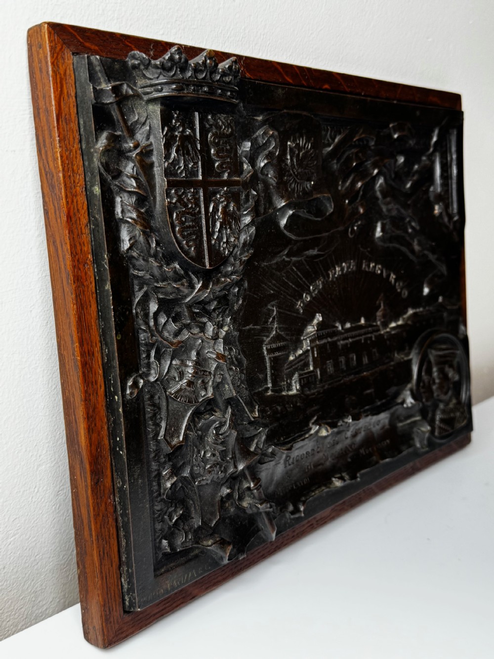 A rectangular dark bronze relief plaque depicting Castello Sforzesco with heraldic coats of arms, mounted inside a wooden frame. View V