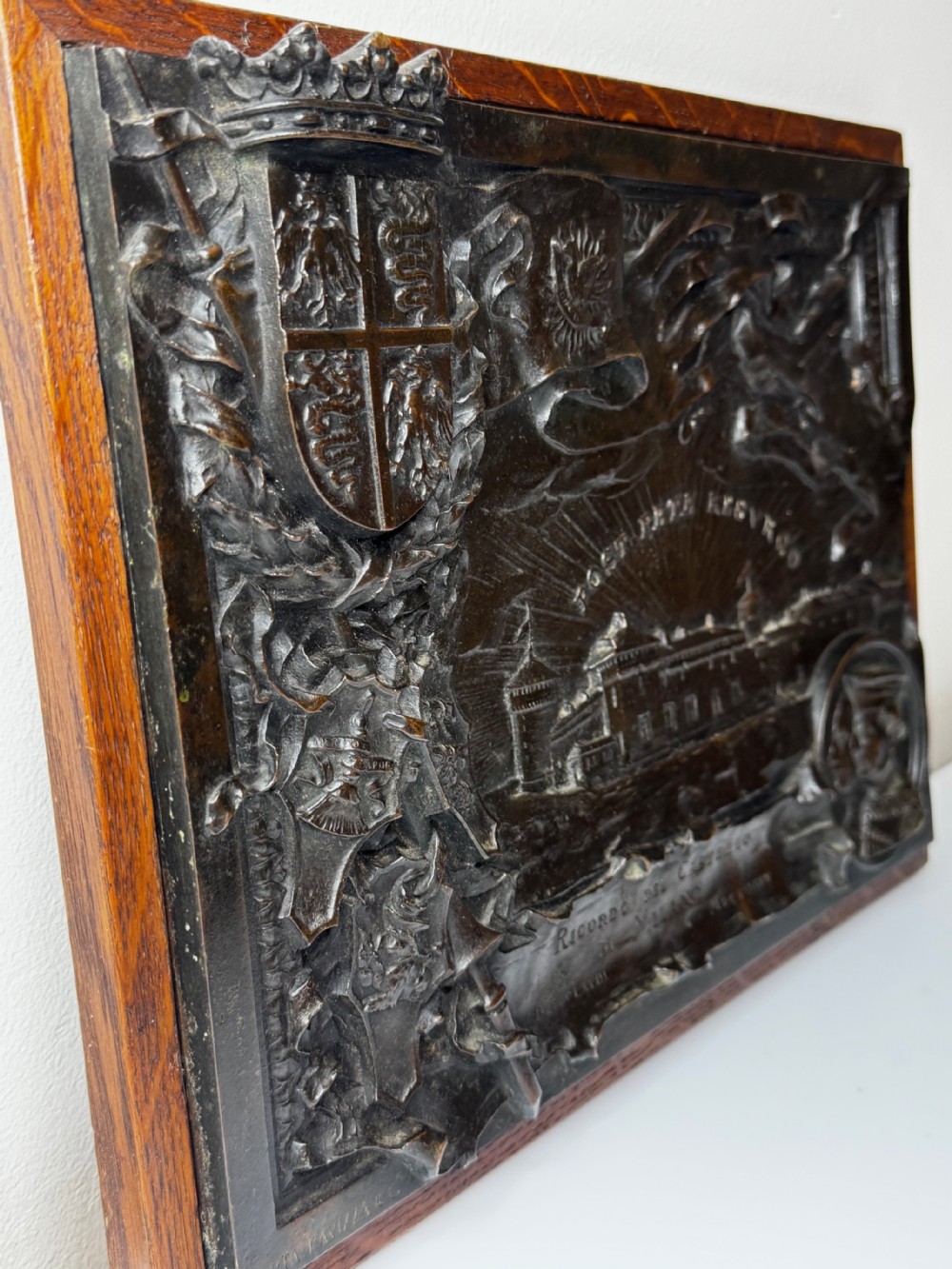 A rectangular dark bronze relief plaque depicting Castello Sforzesco with heraldic coats of arms, mounted inside a wooden frame. View VI