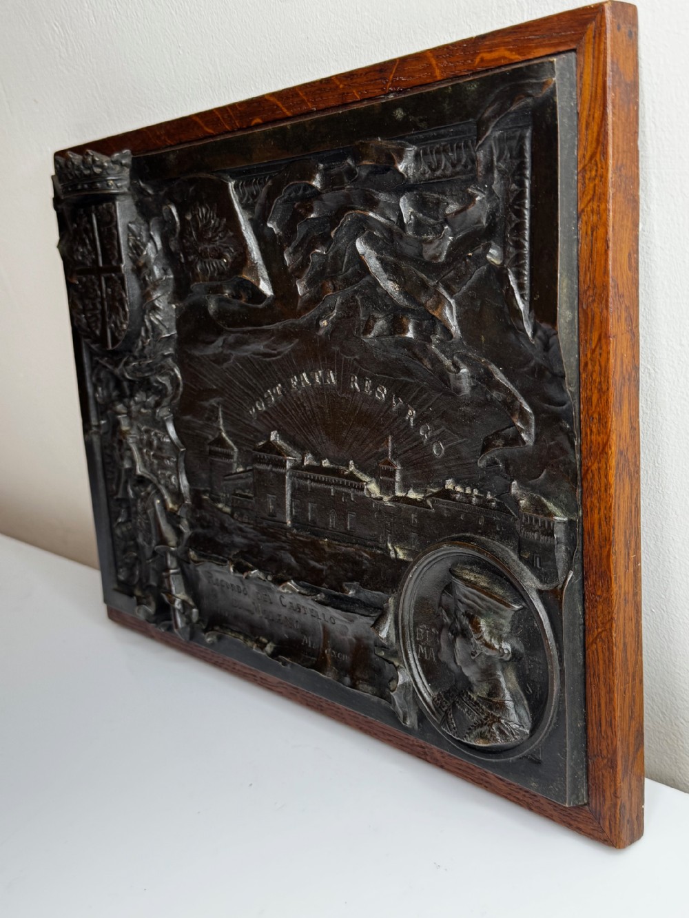 A rectangular dark bronze relief plaque depicting Castello Sforzesco with heraldic coats of arms, mounted inside a wooden frame. View VII
