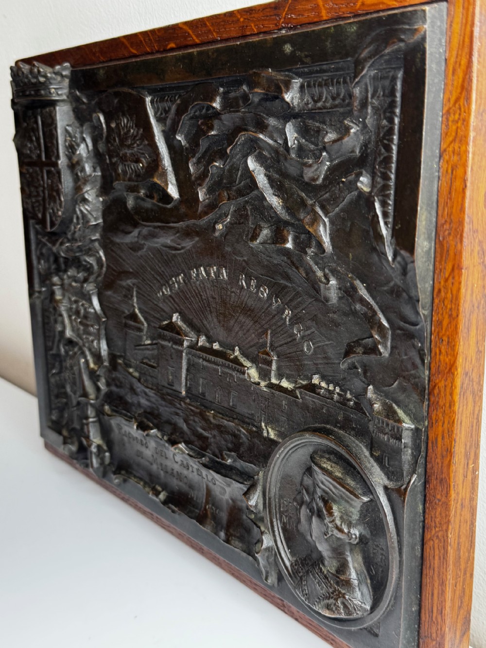 A rectangular dark bronze relief plaque depicting Castello Sforzesco with heraldic coats of arms, mounted inside a wooden frame. View VIII