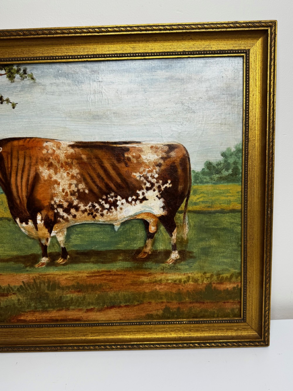 A 19th-century oil painting depicting a brown and white shorthorn bull standing in side profile in a green field, encased in a gilded frame. View VI