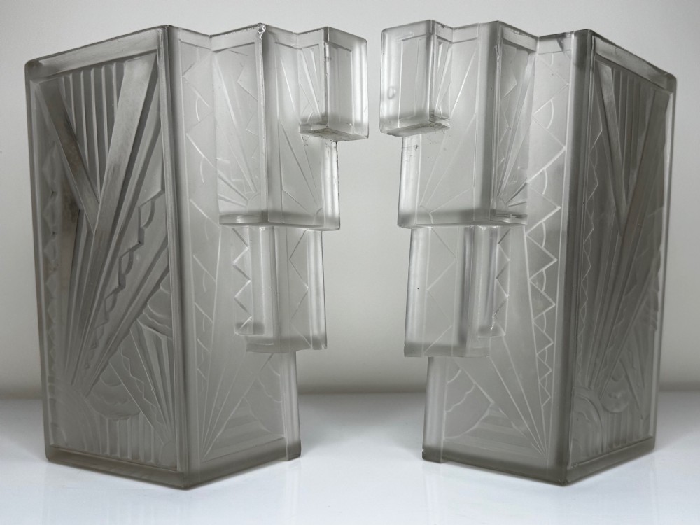 A pair of Art Deco frosted moulded glass wall lights featuring a stepped skyscraper silhouette with geometric sunburst and chevron carvings. View XXX