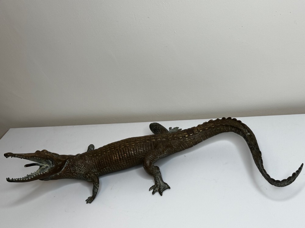 A dark bronze sculpture of a crocodile with an open mouth, detailed scales, and a curved tail designed as a water fountain spout. View II