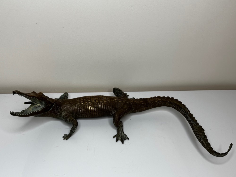 A dark bronze sculpture of a crocodile with an open mouth, detailed scales, and a curved tail designed as a water fountain spout. View III