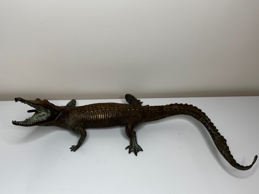 A dark bronze sculpture of a crocodile with an open mouth, detailed scales, and a curved tail designed as a water fountain spout. View IV