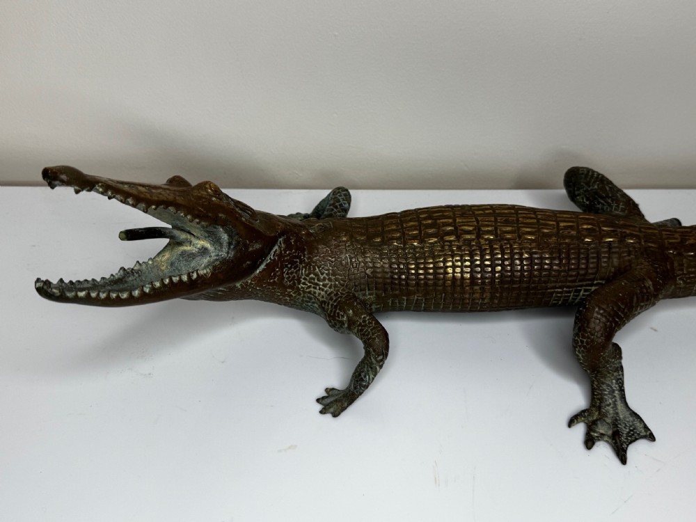 A dark bronze sculpture of a crocodile with an open mouth, detailed scales, and a curved tail designed as a water fountain spout. View VI