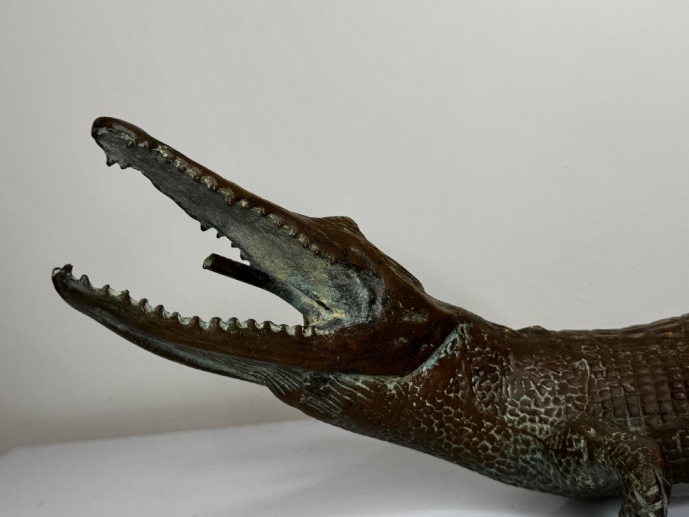 A dark bronze sculpture of a crocodile with an open mouth, detailed scales, and a curved tail designed as a water fountain spout. View XX