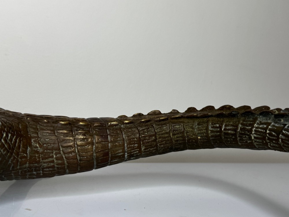 A dark bronze sculpture of a crocodile with an open mouth, detailed scales, and a curved tail designed as a water fountain spout. View XXI