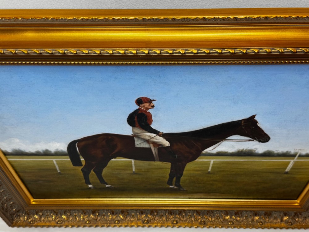 A framed Victorian oil painting depicting a bay racehorse and mounted jockey against a blue sky and green racecourse backdrop. View V