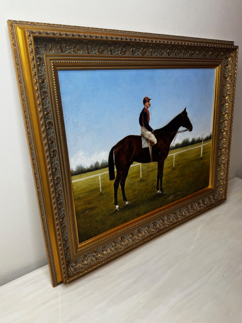 A framed Victorian oil painting depicting a bay racehorse and mounted jockey against a blue sky and green racecourse backdrop. View X