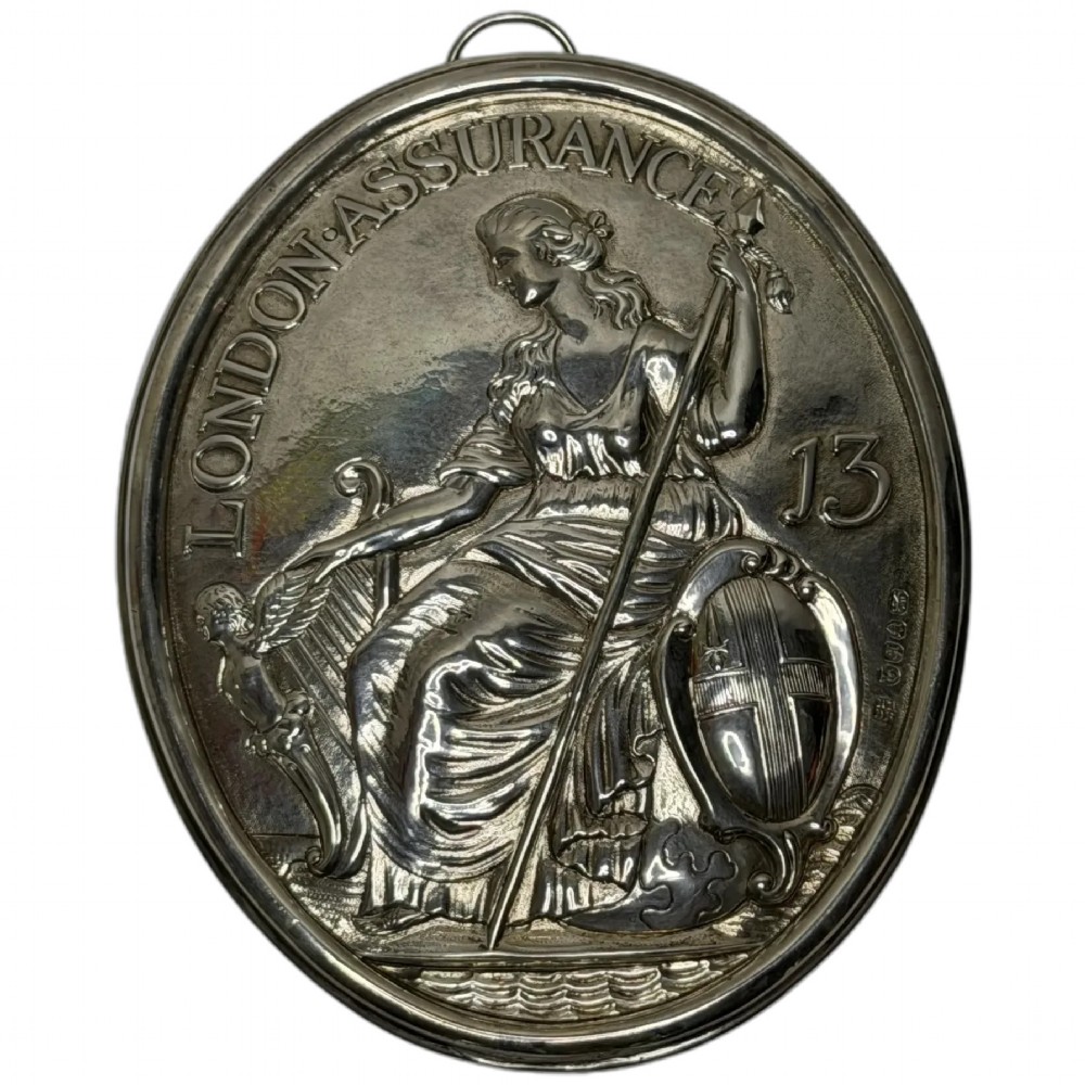 An oval George III sterling silver plaque featuring an embossed relief of Britannia holding a spear beside a shield and harp with embossed text reading London Assurance.