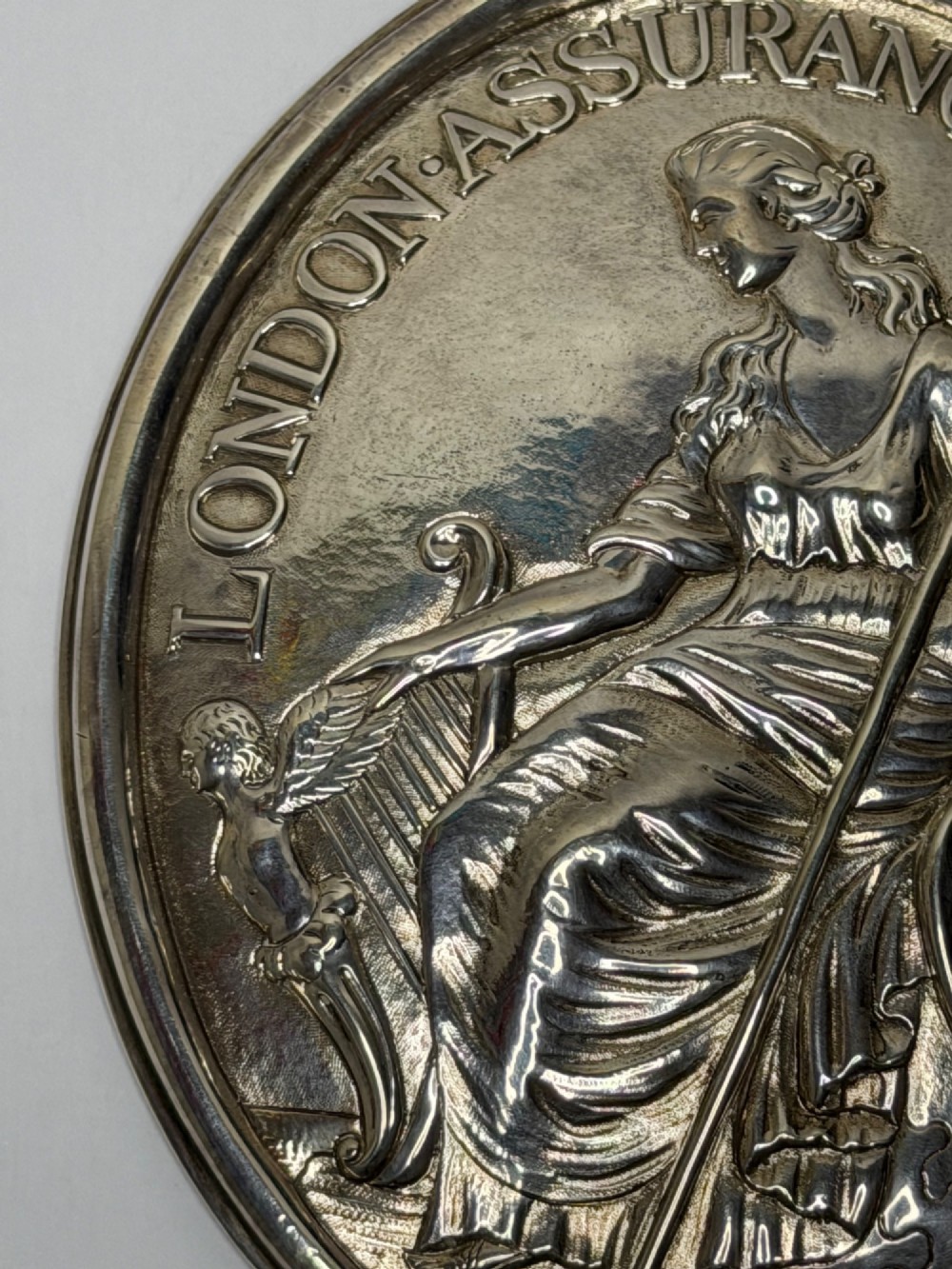 An oval George III sterling silver plaque featuring an embossed relief of Britannia holding a spear beside a shield and harp with embossed text reading London Assurance. View III