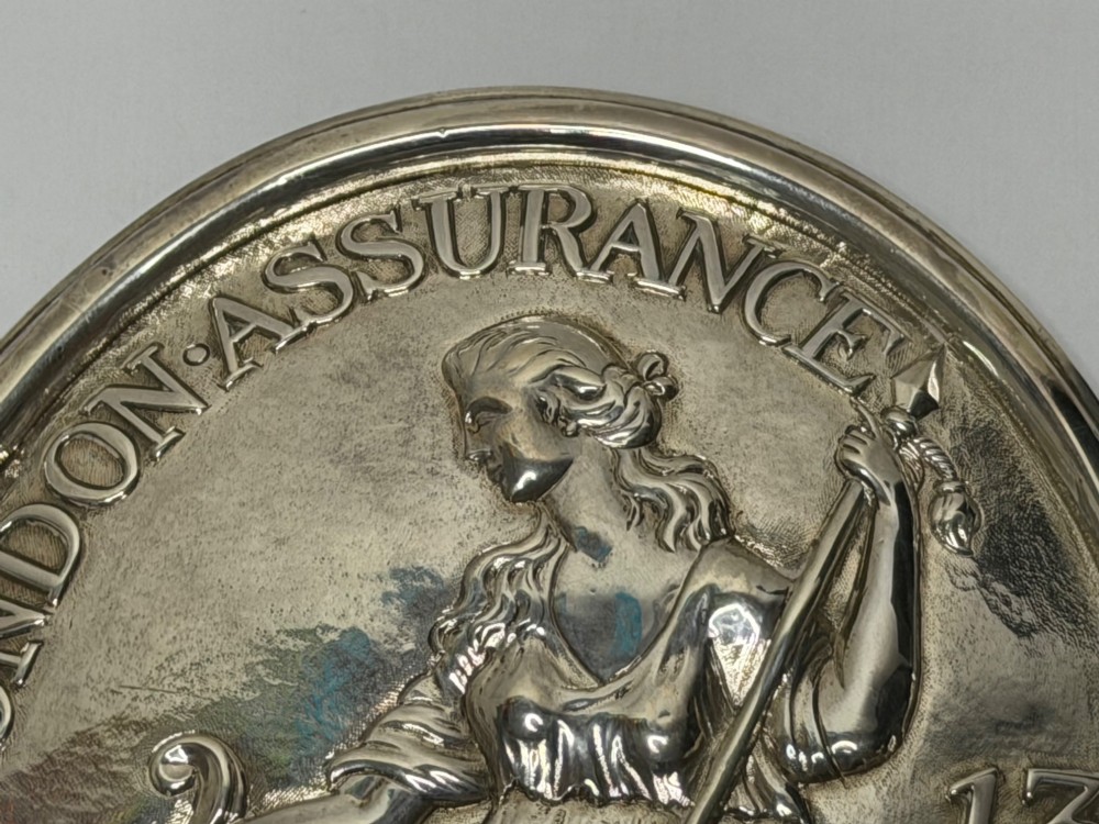 An oval George III sterling silver plaque featuring an embossed relief of Britannia holding a spear beside a shield and harp with embossed text reading London Assurance. View IV