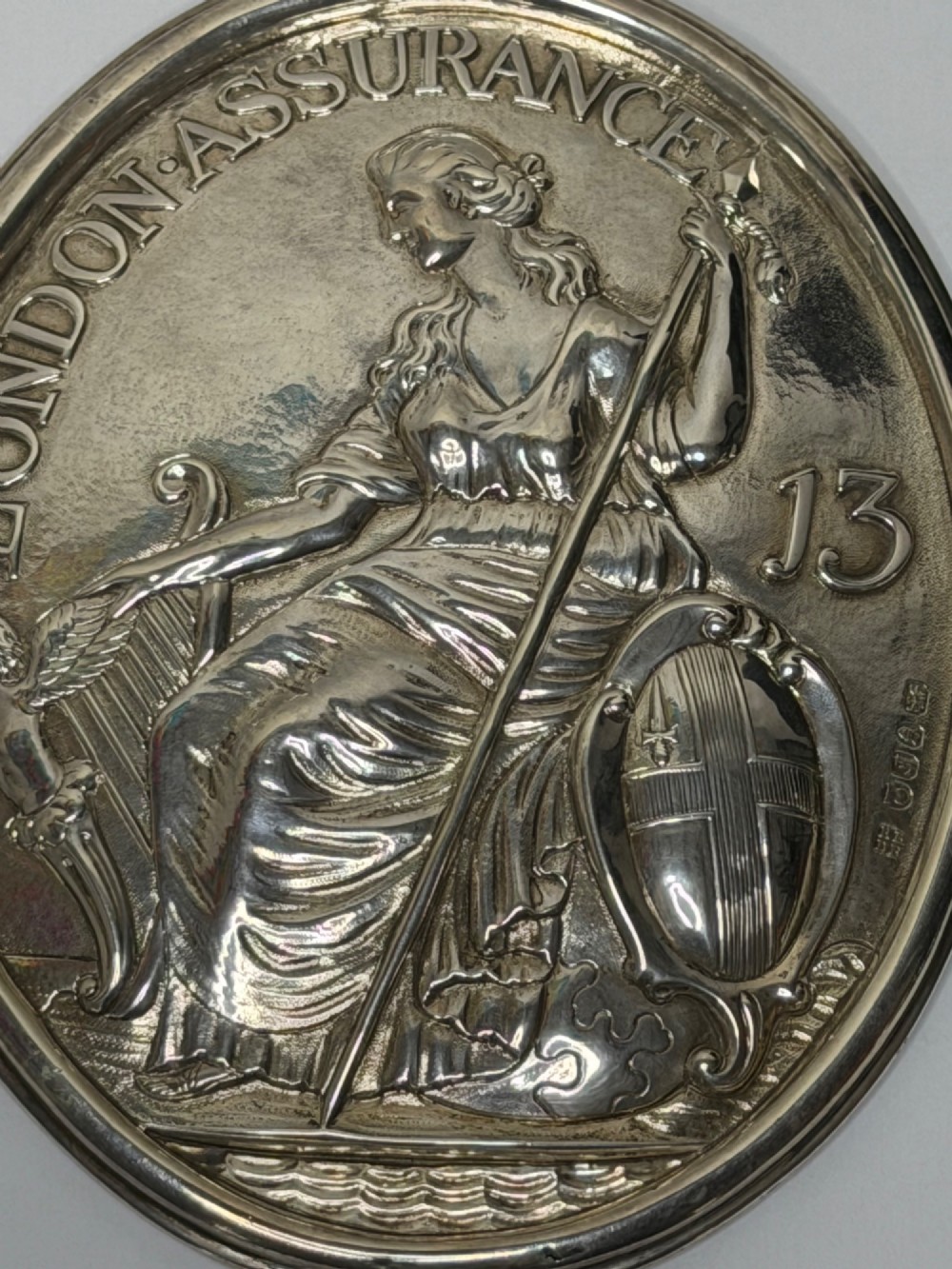 An oval George III sterling silver plaque featuring an embossed relief of Britannia holding a spear beside a shield and harp with embossed text reading London Assurance. View VI