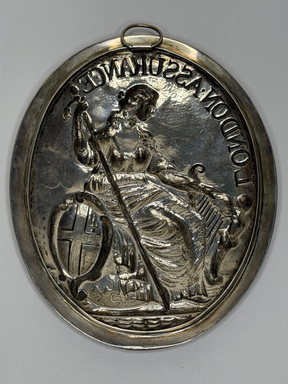 An oval George III sterling silver plaque featuring an embossed relief of Britannia holding a spear beside a shield and harp with embossed text reading London Assurance. View XI
