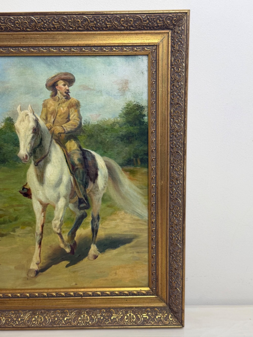 An oil painting depicting a man in a hat and buckskin jacket riding a grey horse along a rural path, set within an ornate gilded wooden frame. View III