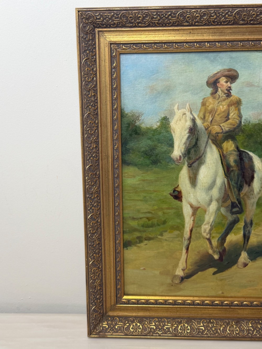 An oil painting depicting a man in a hat and buckskin jacket riding a grey horse along a rural path, set within an ornate gilded wooden frame. View IV