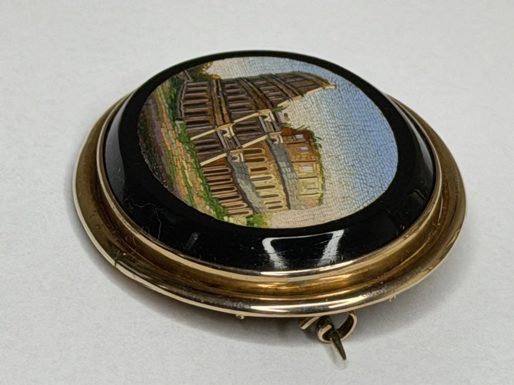 An oval nineteenth-century micromosaic brooch set in a yellow gold bezel depicting the Roman Colosseum against a light blue sky within a black surround. View III