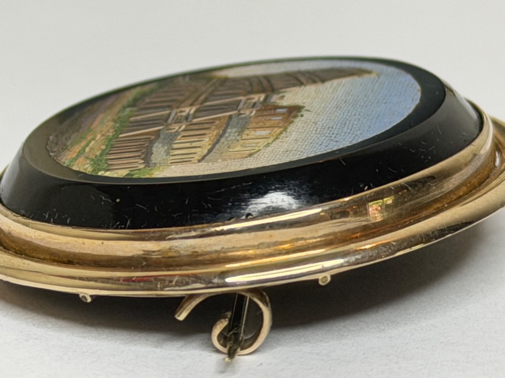 An oval nineteenth-century micromosaic brooch set in a yellow gold bezel depicting the Roman Colosseum against a light blue sky within a black surround. View X