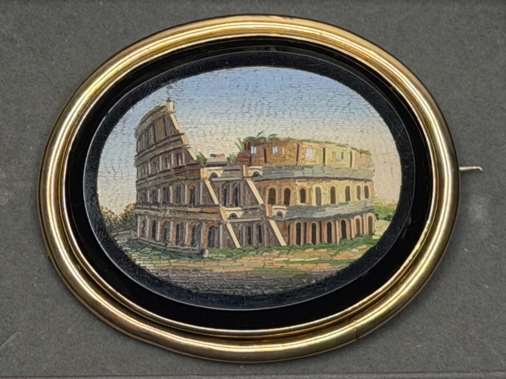 An oval nineteenth-century micromosaic brooch set in a yellow gold bezel depicting the Roman Colosseum against a light blue sky within a black surround. View XIV