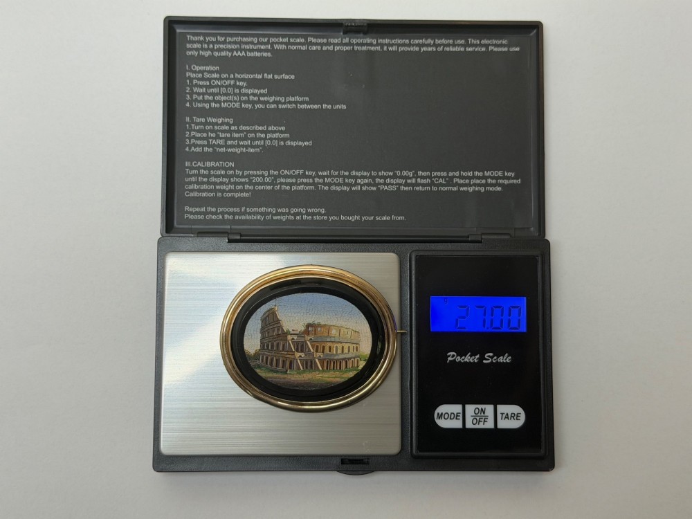 An oval nineteenth-century micromosaic brooch set in a yellow gold bezel depicting the Roman Colosseum against a light blue sky within a black surround. View XV