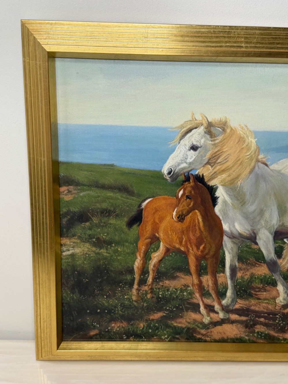 A framed oil painting featuring a grey mare standing beside a young chestnut foal on a grassy coastal headland overlooking the sea. View V