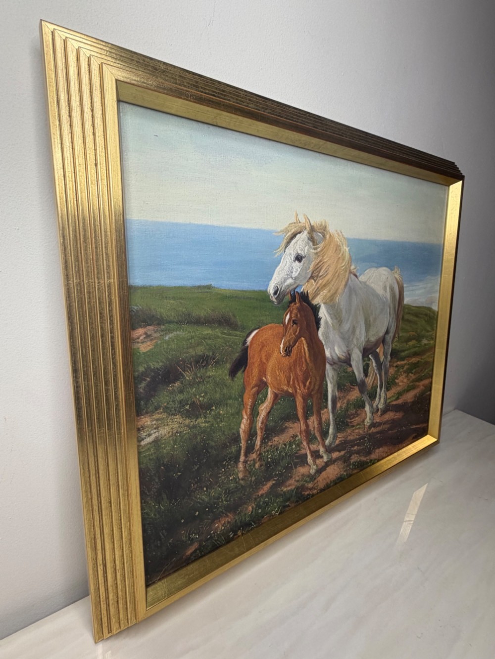 A framed oil painting featuring a grey mare standing beside a young chestnut foal on a grassy coastal headland overlooking the sea. View XV