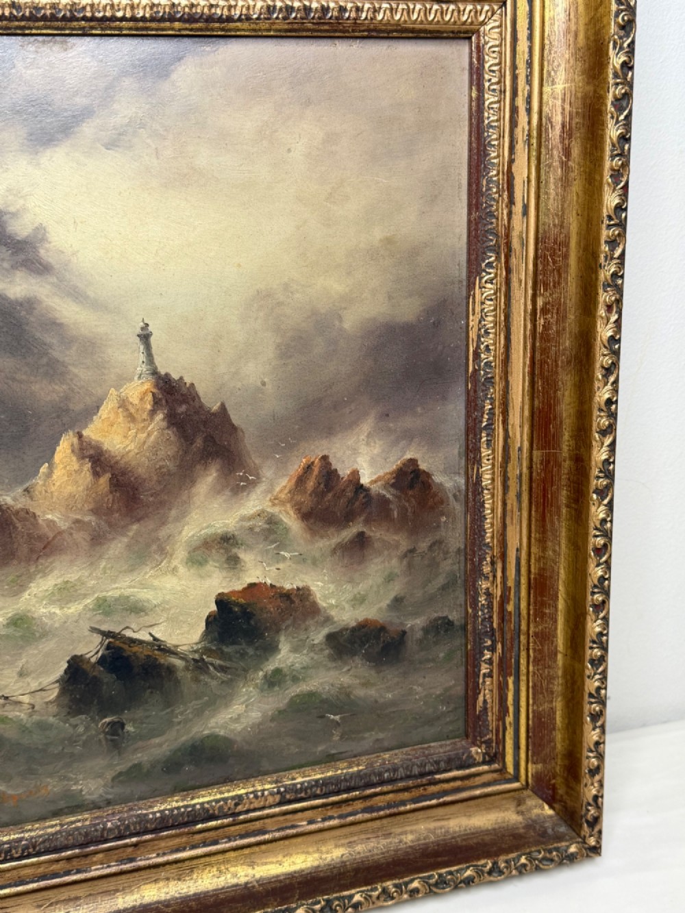 A vertical oil painting framed in carved gold leaf depicting La Corbiere lighthouse atop jagged rocks amidst a stormy grey sea with a shipwreck in the foreground. View III