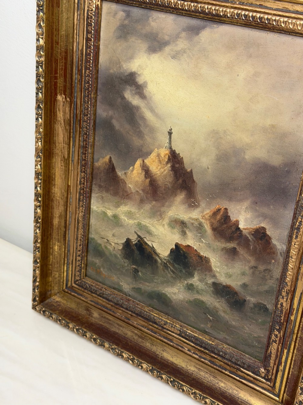 A vertical oil painting framed in carved gold leaf depicting La Corbiere lighthouse atop jagged rocks amidst a stormy grey sea with a shipwreck in the foreground. View IV