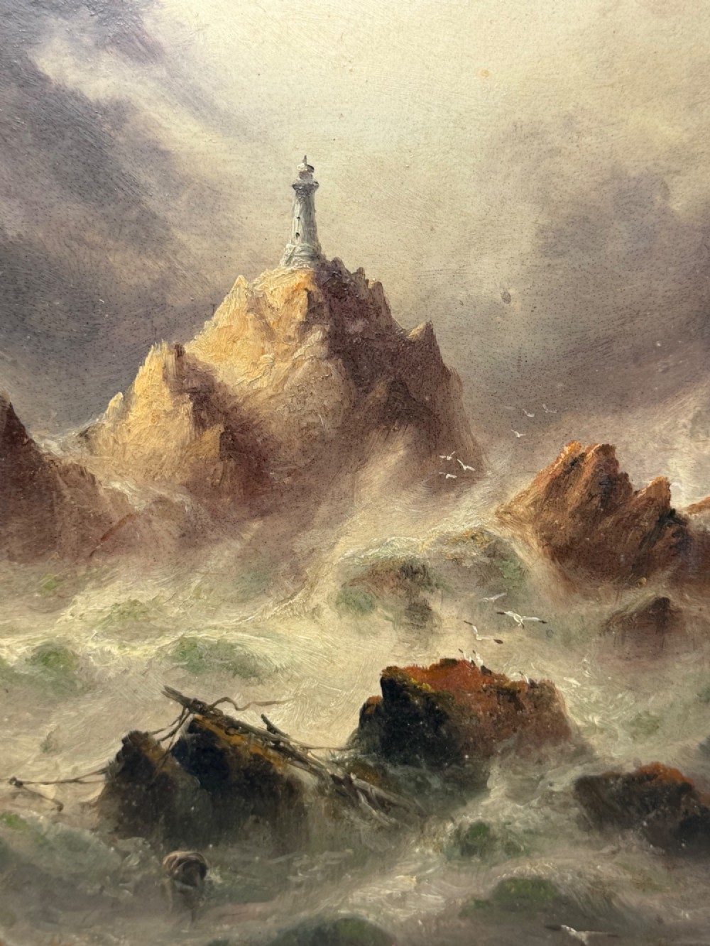 A vertical oil painting framed in carved gold leaf depicting La Corbiere lighthouse atop jagged rocks amidst a stormy grey sea with a shipwreck in the foreground. View VI