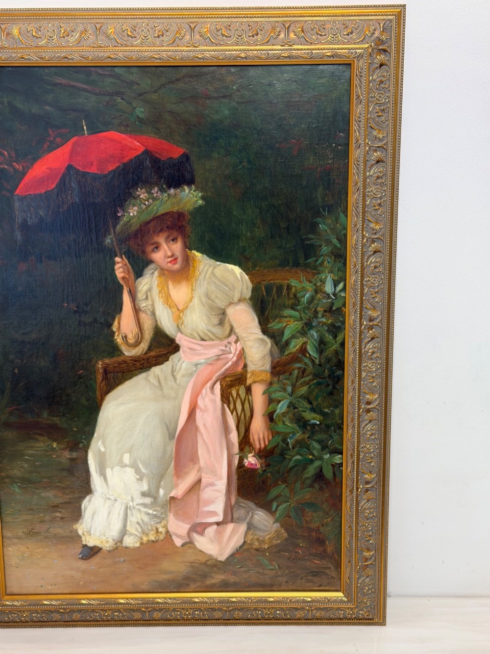 An antique oil painting depicting a seated woman in a white dress holding a red parasol, presented in an ornate carved gilded frame. View III