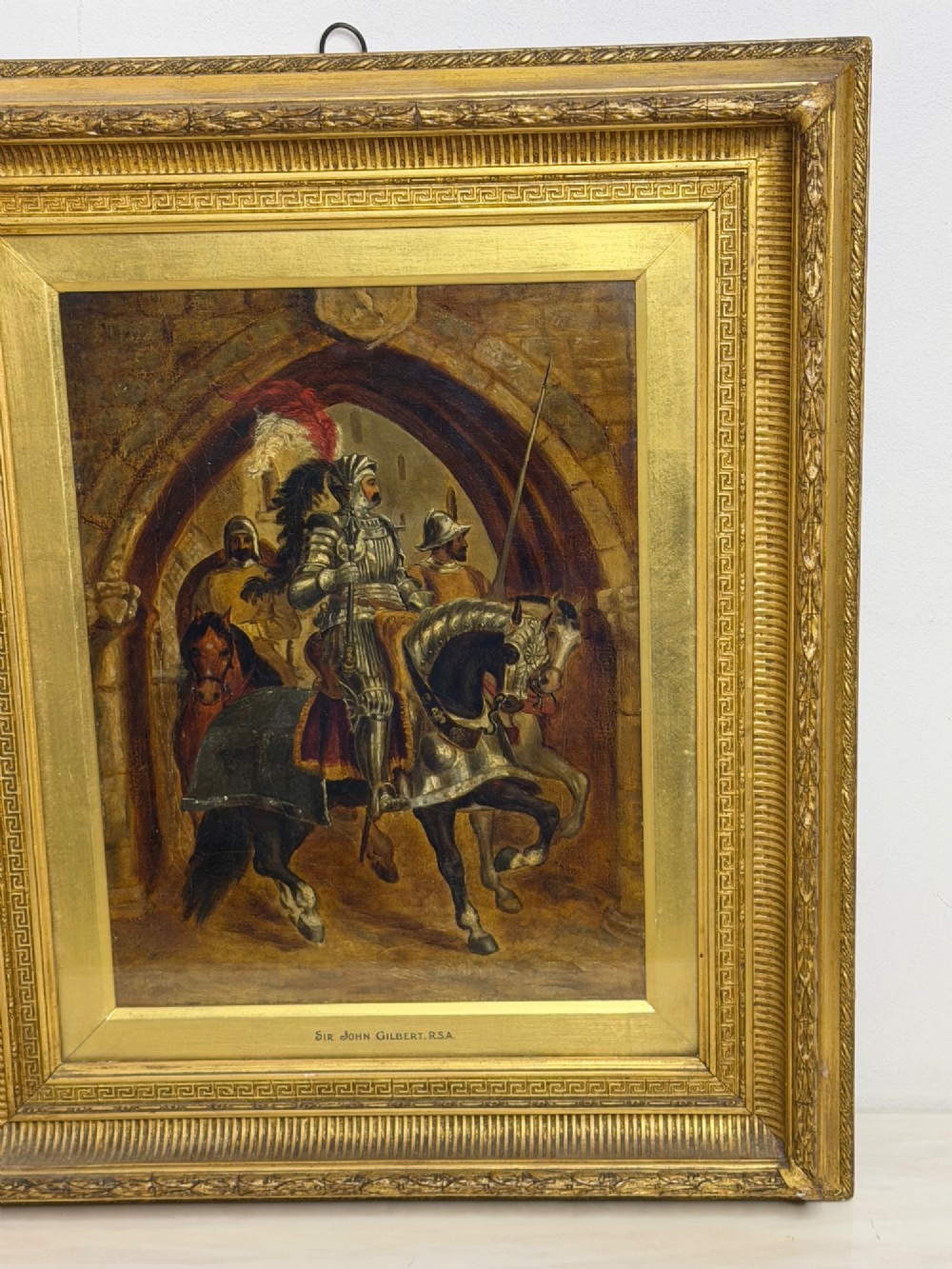 An antique oil painting depicting a knight in full armour riding a horse beneath a stone archway, framed in an ornate gilded wood frame. View II