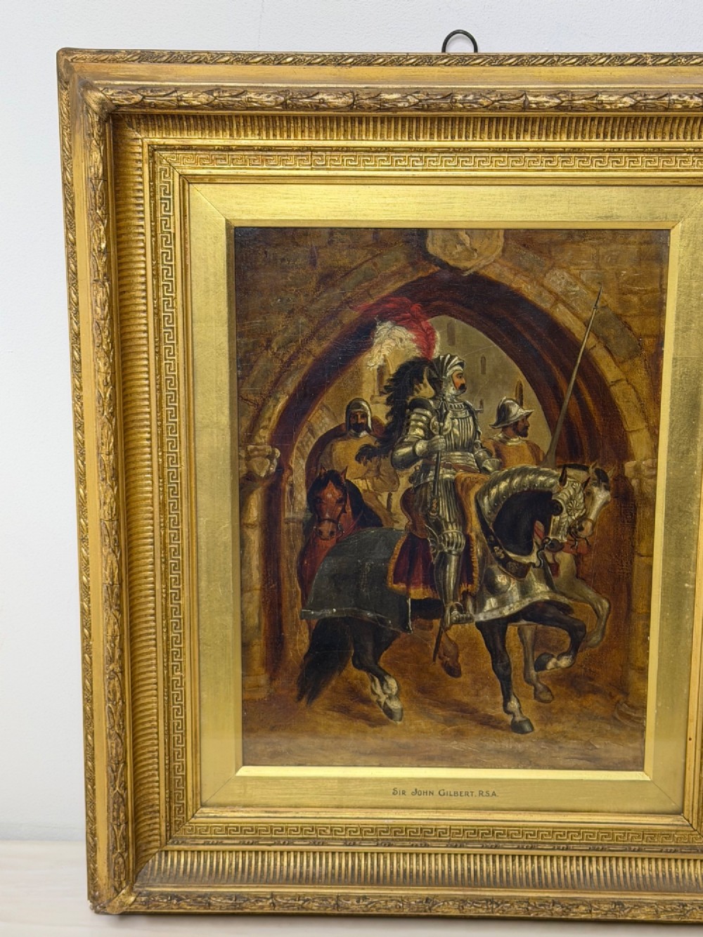 An antique oil painting depicting a knight in full armour riding a horse beneath a stone archway, framed in an ornate gilded wood frame. View III