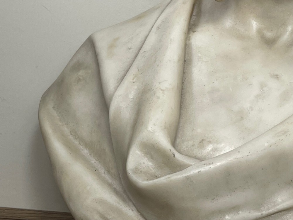 A carved off-white marble bust depicting a bald male figure with a prominent moustache wearing classical draped fabric across the shoulders. View V