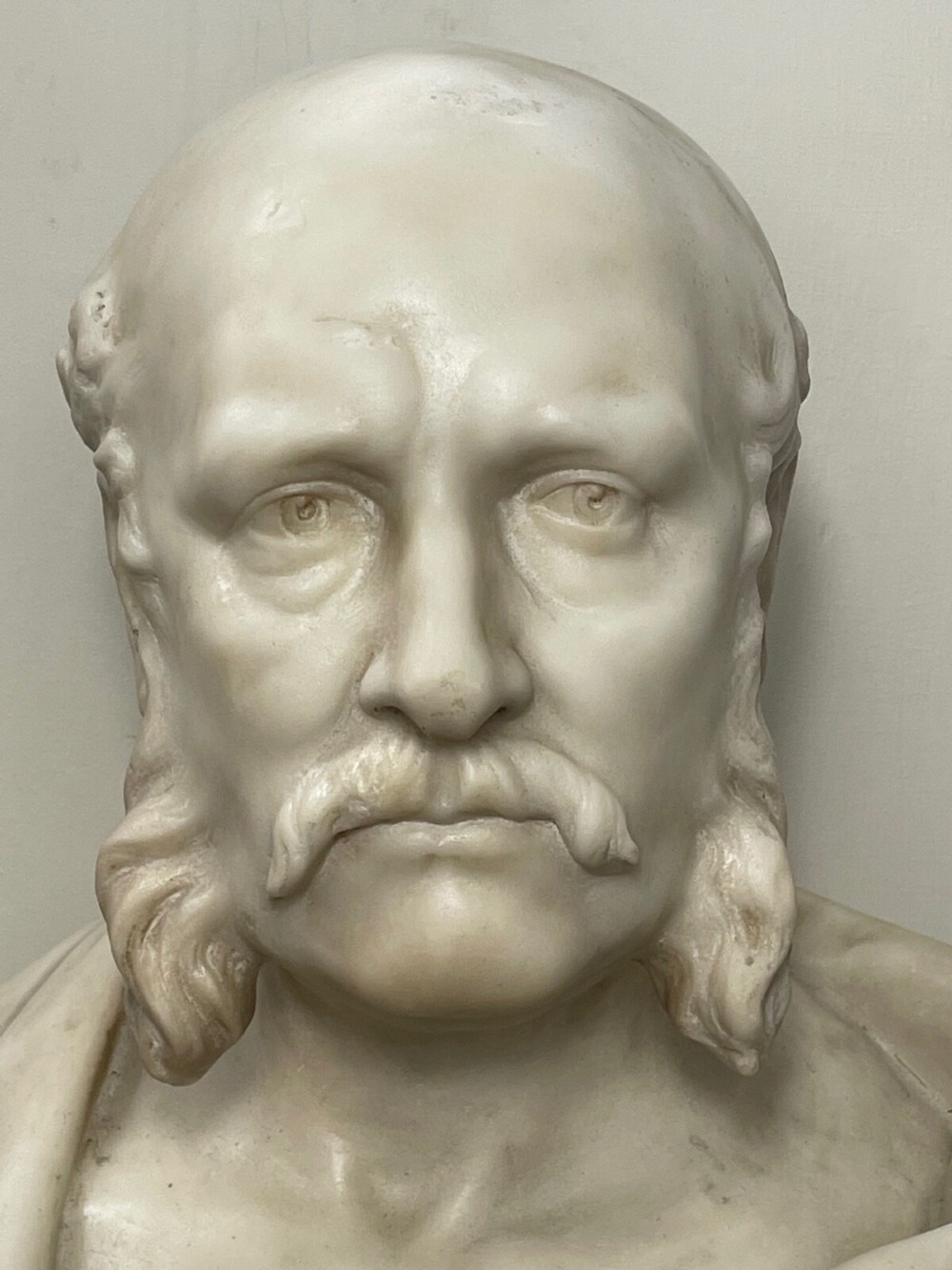 A carved off-white marble bust depicting a bald male figure with a prominent moustache wearing classical draped fabric across the shoulders. View VI