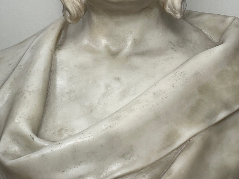 A carved off-white marble bust depicting a bald male figure with a prominent moustache wearing classical draped fabric across the shoulders. View VII