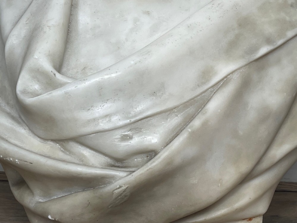 A carved off-white marble bust depicting a bald male figure with a prominent moustache wearing classical draped fabric across the shoulders. View VIII
