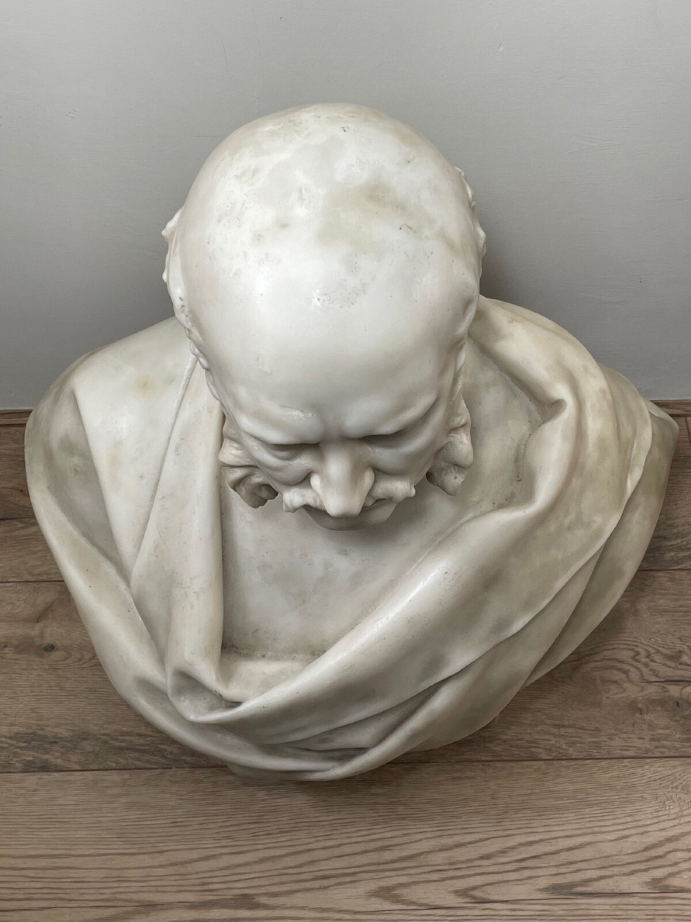 A carved off-white marble bust depicting a bald male figure with a prominent moustache wearing classical draped fabric across the shoulders. View IX