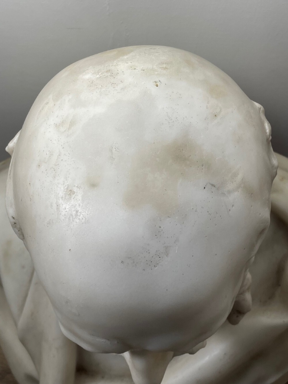 A carved off-white marble bust depicting a bald male figure with a prominent moustache wearing classical draped fabric across the shoulders. View X