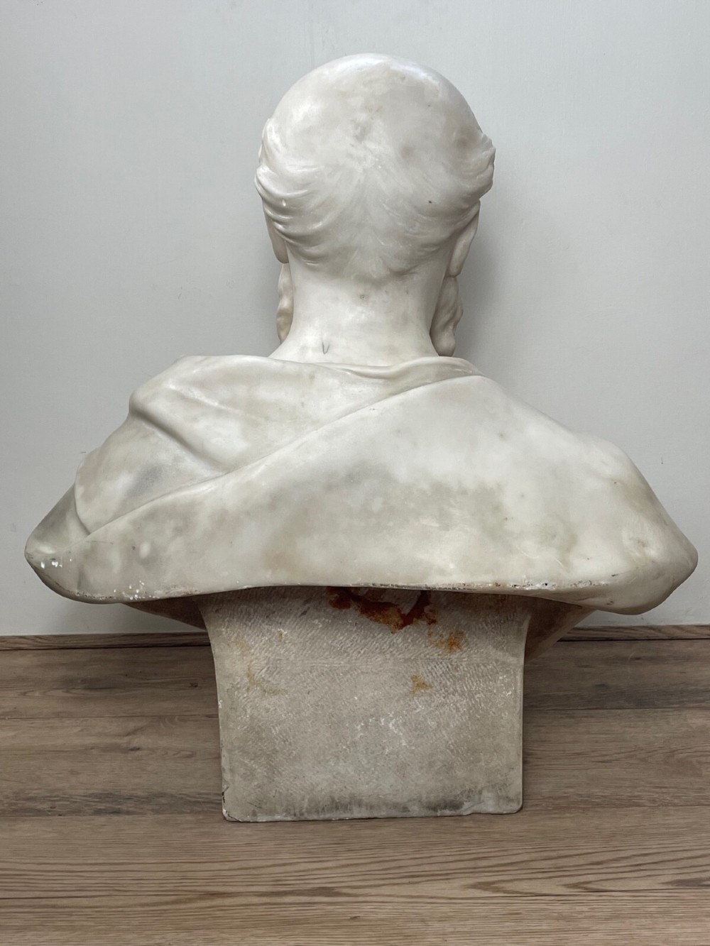 A carved off-white marble bust depicting a bald male figure with a prominent moustache wearing classical draped fabric across the shoulders. View XII