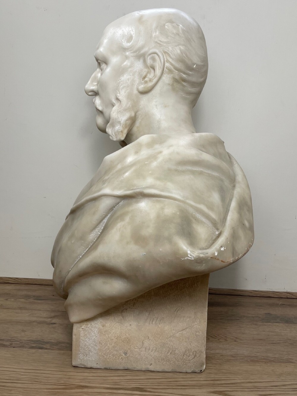 A carved off-white marble bust depicting a bald male figure with a prominent moustache wearing classical draped fabric across the shoulders. View XV